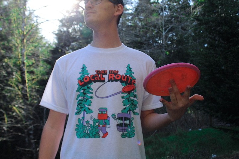 May include: A white t-shirt with a graphic of a cartoon character holding a disc golf basket. The graphic says "Take the Local Route". The person is holding a pink disc golf disc.