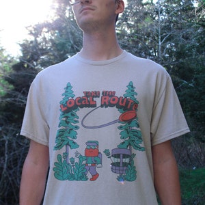 May include: A beige t-shirt with a graphic of a person throwing a disc in a forest. The graphic says "Take the Local Route".