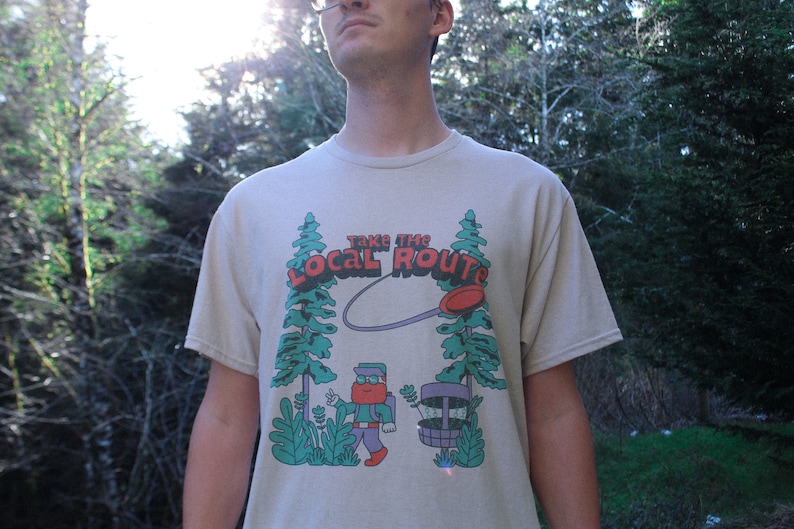 May include: A beige t-shirt with a graphic of a person throwing a disc in a forest. The graphic says "Take the Local Route".
