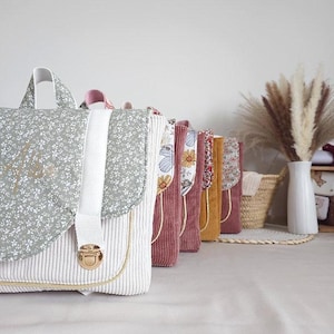 Puede incluir: Una fila de mochilas de pana en varios colores, incluyendo verde salvia, rosa empolvado, amarillo mostaza y malva. Cada mochila cuenta con una solapa con estampado floral, una correa vertical blanca y un cierre de metal dorado al frente.