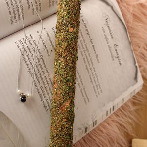 Könnte beinhalten: Eine grüne und braune Kräuterkerze mit goldenen Flocken. Die Kerze liegt auf einem Buch mit deutschem Text über negative Energien und wie man sich vor dem bösen Blick schützen kann.