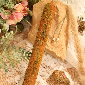 May include: A long, brown candle with green herbs and spices embedded in the wax. The candle is sitting on a rock with a green string necklace with white beads.