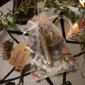 Könnte beinhalten: Ein durchsichtiger Netzbeutel gefüllt mit kleinen Steinen, einer Kerze und einem goldenen Stab. Ein kleines Etikett trägt die Aufschrift "Mystery Sachet". Die Artikel sind auf einem silbernen Tablett mit schwarzen Stern- und Kartendesigns und Zweigen angeordnet.