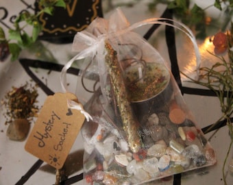 Può includere: Un sacchetto di rete trasparente pieno di piccole pietre, una candela e un bastoncino dorato. Un piccolo cartellino recita "Mystery Sachet". Gli oggetti sono disposti su un vassoio d'argento con disegni di stelle e carte nere e rametti di verde.