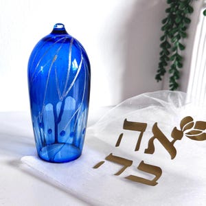 Peut inclure: Un dôme en verre bleu cobalt orné de fines lignes gravées argentées et d'un sommet arrondi. À côté de l'objet en verre se trouve une pochette en tissu blanc transparent décorée d'inscriptions en hébreu dorées et d'un motif floral.
