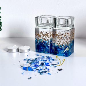 May include: Two tealight candle holders with gold leaf accents and blue and green glass shards embedded in the resin. The holders are sitting on a white surface with some of the glass shards scattered around them.