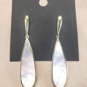 Pode incluir: Um par de brincos pendurados dourados. Cada brinco apresenta uma incrustação perolada em forma de gota, emoldurada por uma fina borda dourada. Os brincos são exibidos em um cartão preto com um furo na parte superior.