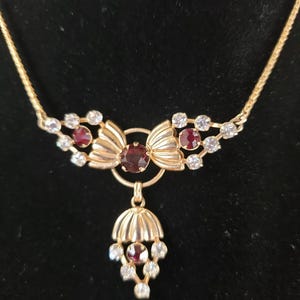 May include: Gold-toned necklace with a pendant featuring a cluster of red and clear gemstones. The necklace has a delicate chain and a decorative bow-shaped centerpiece. The pendant has a teardrop shape with a cluster of gemstones.