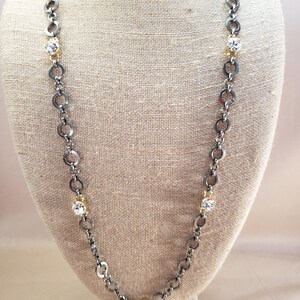 May include: A long necklace with a gunmetal chain featuring alternating round links and small gold-toned accents. The necklace has clear, round crystal accents. The necklace is displayed on a mannequin.