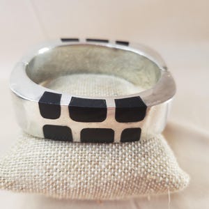 May include: A silver-toned square bangle bracelet with black rectangular inlays. The bracelet is displayed on a small, textured, beige pillow. The jewelry piece has a modern, geometric design.