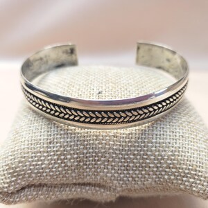 May include: A silver cuff bracelet featuring a central band with a detailed, repeating braided or wheat-like pattern. The metal has a polished finish with darkened recessed areas that highlight the intricate texture of the design along the center.