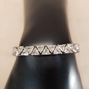 May include: A gold-toned bracelet featuring a row of clear, triangular-cut gemstones. The gemstones are set closely together, creating a continuous sparkle. The bracelet is displayed on a black surface, highlighting the jewelry's brilliance.