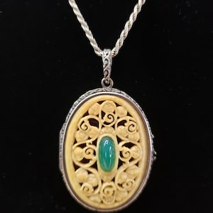 Puede incluir: Un colgante de medallón ovalado en una cadena de cuerda plateada. El medallón presenta un intrincado diseño de filigrana floral tallada en color crema que rodea una piedra preciosa verde ovalada, lisa y pulida. El marco de metal tiene un acabado plateado.