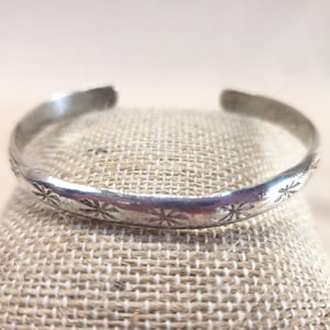 Puede incluir: Un brazalete plateado con un patrón floral. El brazalete abierto se muestra sobre una superficie beige. El brazalete tiene una serie de pequeños diseños en forma de estrella grabados alrededor de la banda.