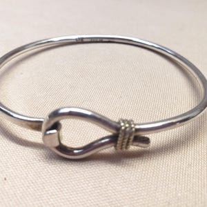 May include: A polished silver bangle bracelet featuring a central knot-like design. The metal band is smooth and circular, with a textured, rope-like detail wrapping around the point where the ends meet. The hallmark 925 is visible on the band.