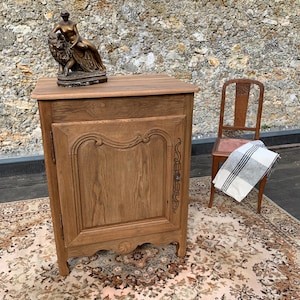 Peut inclure: Un buffet en bois brun clair avec une seule porte et des sculptures décoratives. Une statue en bronze d'une femme et d'un lion est posée dessus. Un tapis à motifs et une chaise en bois sont en arrière-plan.