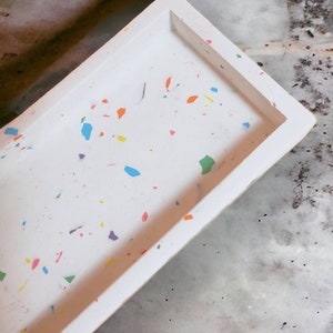 Op de afbeelding: Een wit rechthoekig dienblad met een gespikkeld patroon van kleurrijke verfspatten. Het dienblad is gemaakt van een glad, duurzaam materiaal en heeft een licht verhoogde rand.