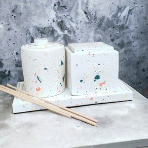 Op de afbeelding: Twee witte keramische containers met een gespikkeld patroon in tinten oranje, groen en geel. De containers staan op een witte dienblad met een gespikkeld patroon. Er liggen vier houten stokjes op het dienblad.