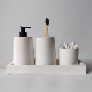 Puede incluir: Bandeja organizadora de baño de cerámica blanca con tres compartimentos. La bandeja contiene una botella de bomba blanca, un portacepillos de dientes blanco con un cepillo de dientes de madera y un portaalgodón blanco.