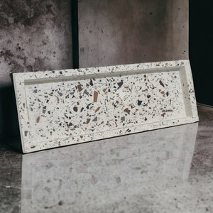 May include: A white rectangular tray with a speckled terrazzo design. The tray is made of concrete and has a smooth, polished finish.