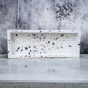 May include: A white rectangular tray with a speckled black and gray terrazzo pattern. The tray is made of concrete and has a smooth, polished finish.