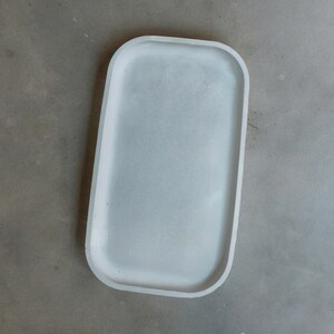 Può includere: Un vassoio rettangolare bianco con bordi arrotondati. Il vassoio è in cemento e ha una finitura liscia e opaca.