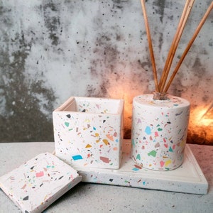 Op de afbeelding: Een witte terrazzo set met een vierkante container, een cilindrische diffuser met stokjes en een kleine vierkante onderzetter. De set heeft kleurrijke vlekken van blauw, geel, roze en groen. De items zijn gerangschikt op een rechthoekige tray.