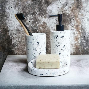 Puede incluir: Un juego de baño blanco con motas negras. El juego incluye un portacepillos de dientes, un dispensador de jabón y un jabonera. La jabonera tiene una pastilla de jabón.