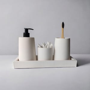 May include: White ceramic bathroom countertop organizer with three compartments. The organizer includes a pump bottle, a cotton swab holder, and a toothbrush holder. The organizer is on a white tray.
