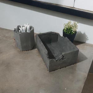 Puede incluir: Dos contenedores de hormigón, uno con un exterior rugoso y texturizado y el otro con un exterior liso y texturizado. El contenedor con el exterior rugoso está lleno de bastoncillos de algodón. El contenedor con el exterior liso está vacío.