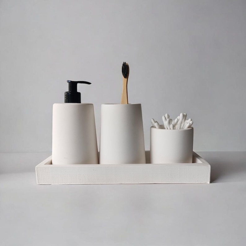 May include: White ceramic bathroom organizer tray with three compartments. The tray holds a white pump bottle, a white toothbrush holder with a wooden toothbrush, and a white cotton swab holder.