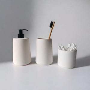 May include: Three white ceramic bathroom containers. The container on the left is a pump bottle, the middle container is a toothbrush holder, and the container on the right holds cotton swabs.