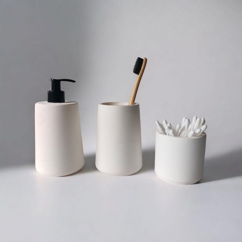 May include: Three white ceramic bathroom containers. The container on the left is a pump bottle, the middle container is a toothbrush holder, and the container on the right holds cotton swabs.