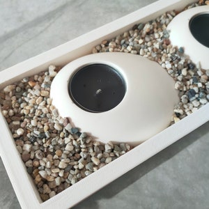 Puede incluir: Portavelas de cerámica blanca con velas negras, colocados en una bandeja blanca llena de pequeñas piedras grises y blancas.