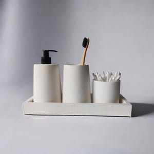 May include: White ceramic bathroom countertop organizer with a tray, two cups, and a pump bottle. The organizer includes a toothbrush and cotton swabs.