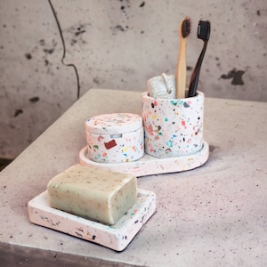 Puede incluir: Un juego de baño con un jabonera con jabón, un pequeño recipiente con tapa y un porta cepillos de dientes. El juego es blanco con motas coloridas de estilo terrazo. Dos cepillos de dientes y un paño están en el soporte.