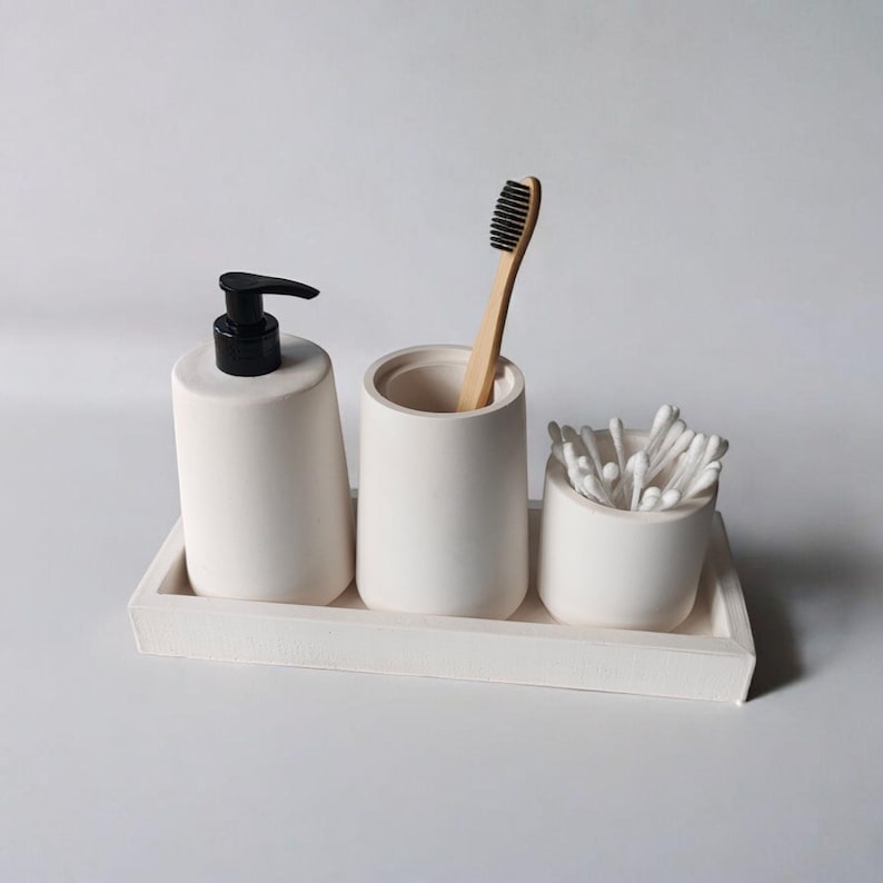 May include: White ceramic bathroom countertop organizer with three compartments. The organizer includes a soap dispenser, a toothbrush holder, and a cotton swab holder.