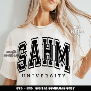 May include: A white t-shirt with a black graphic that says "SAHM UNIVERSITY" in a distressed, vintage-style font. The text is in a large, bold font and has a black outline. The graphic is centered on the shirt and is surrounded by a white background.