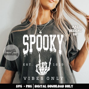 May include: A gray t-shirt with a white graphic design that says "SPOOKY" with dripping paint, a skeleton hand giving the middle finger, "EST. 1629", and "VIBES ONLY".