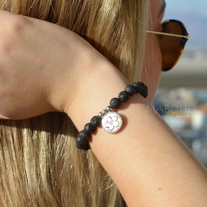 May include: A black lava rock bracelet with a silver flower charm. The bracelet is on a person's wrist.