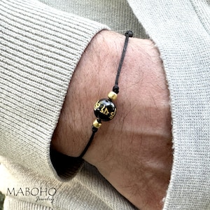 May include: A black string bracelet with a single black bead. The bead has gold lettering on it.