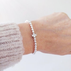 May include: Silver beaded bracelet with a white cross charm. The bracelet is made of small, round silver beads and features a single cross charm in the center. The bracelet is worn on a wrist, with a light-colored sweater sleeve visible.