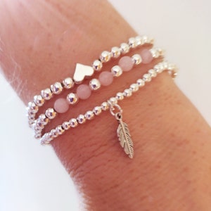 May include: Three silver beaded bracelets with a pink stone charm and a silver feather charm. The bracelets are stacked on a person's wrist.