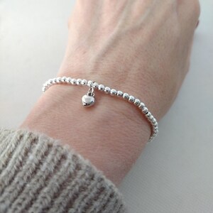 May include: A silver beaded bracelet featuring a small heart pendant. The bracelet is composed of small, spherical beads. The heart charm is suspended from the centre. The wrist is wearing a beige jumper.