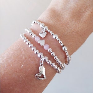 May include: Three silver bracelets with heart charms and pink beads. The bracelets are stacked on a person's wrist.