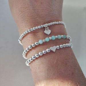 May include: Three beaded bracelets. Two are silver with small round beads, one with a heart charm. The other bracelet has small, round, light blue beads. The bracelets are stacked on a wrist.