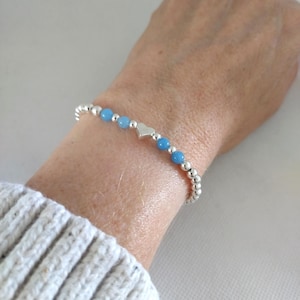 May include: A bracelet with small silver beads and light blue beads, and a silver heart charm. The bracelet is worn on a wrist. The background includes a white sweater.
