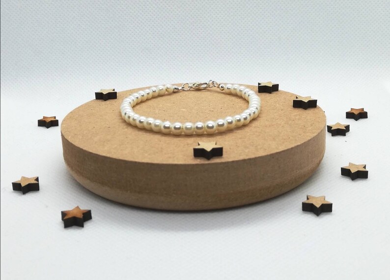 May include: A white pearl bracelet with a silver clasp. The bracelet is on a wooden surface with small wooden stars scattered around it.