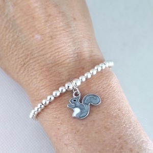 May include: A silver beaded bracelet featuring a grey and white squirrel charm. The charm is an enamel design. The bracelet is worn on a wrist.