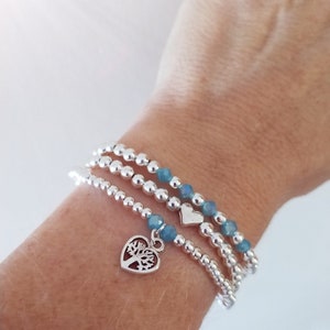 May include: A set of three silver bead bracelets with blue beads. The bottom bracelet has a silver charm in the shape of a heart with a tree inside.